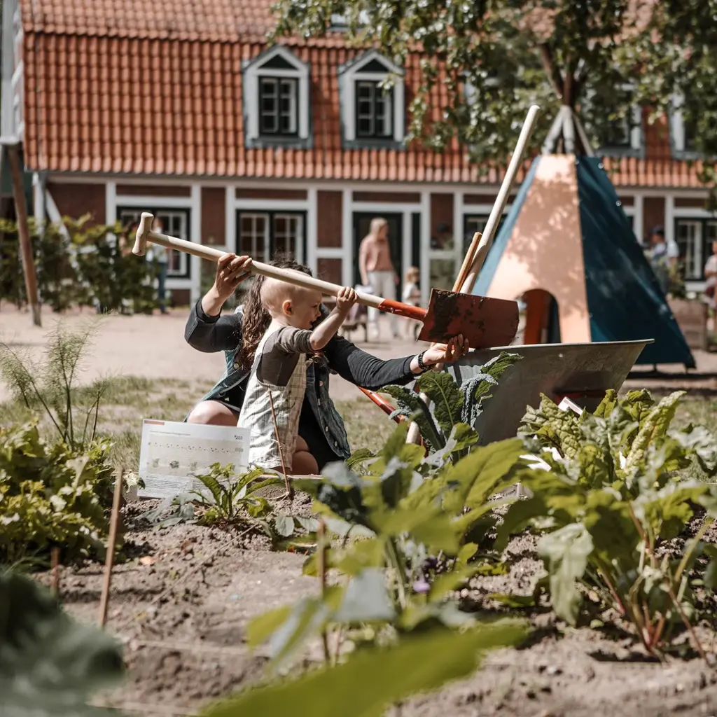 Kind und Erzieherin arbeiten gemeinsam im Gemüsegarten vor einem Kita-Gebäude
