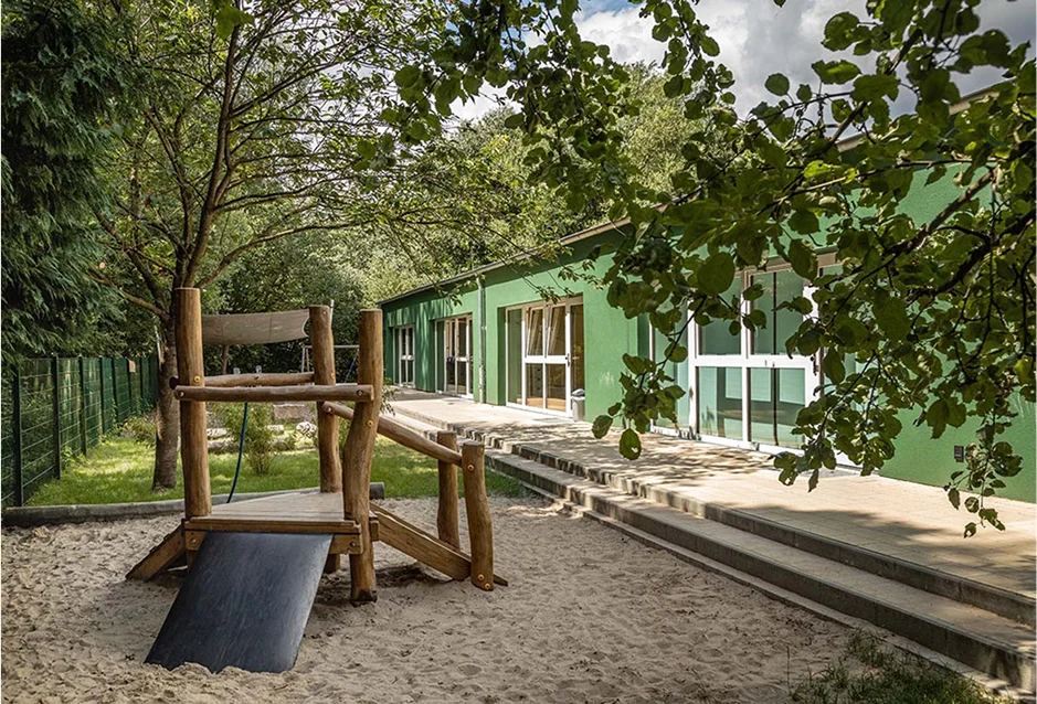 Grünes Kita-Gebäude mit Sandspielplatz und Holzspielgerät im Schatten der Bäume