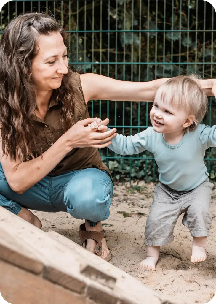 Erzieherin unterstützt Kleinkind beim Laufen im Sandspielbereich